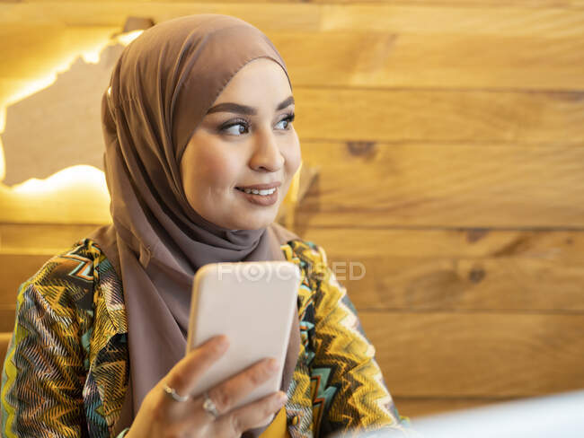 Smiling woman wearing hijab looking away while using mobile phone at cafe — Stock Photo