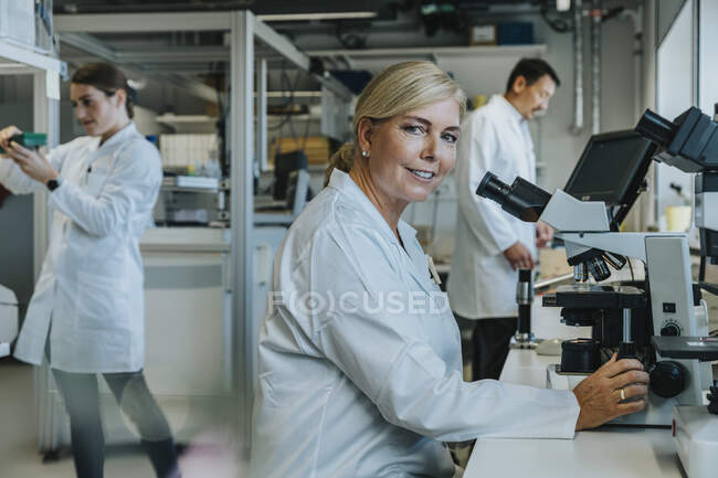 Científico sonriente sentado al microscopio con compañeros de trabajo trabajando en el laboratorio - foto de stock