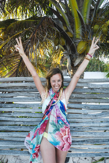 Cheerful woman peace gesturing with arms raised against wooden fence at beach — Stock Photo