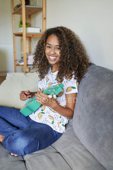 Lächelnde junge Afro-Frau mit Ukulele sitzt auf Sofa im Wohnzimmer — Stockfoto