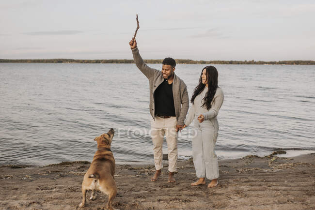 Couple tenant la main tout en jouant avec le chien contre le lac pendant le coucher du soleil — Photo de stock