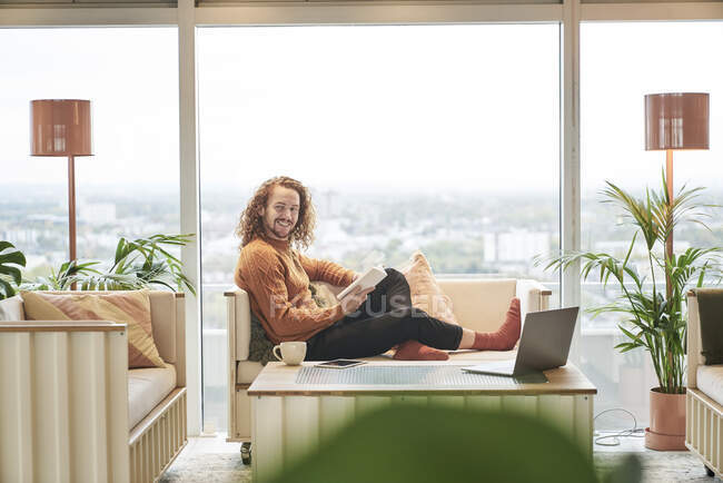 Хипстер улыбается, читая книгу, сидя на диване в гостиной — стоковое фото