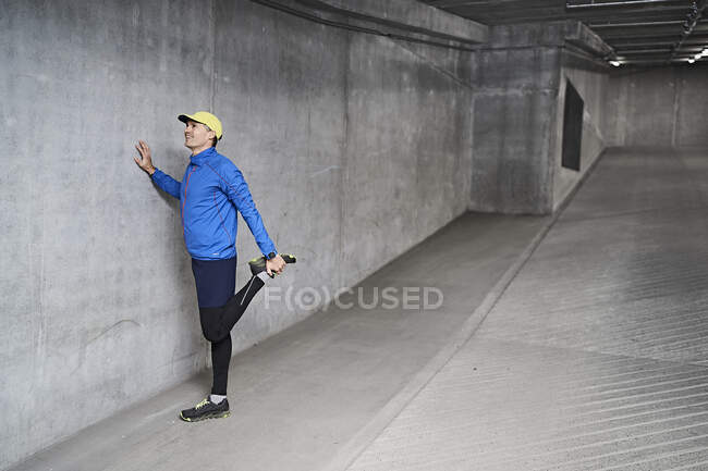 Sourire homme mûr étirement jambe par mur gris dans le garage de stationnement — Photo de stock