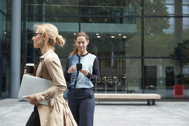 Жінка тримає ноутбук під час ходьби з діловою жінкою за допомогою мобільного телефону на задньому плані — стокове фото