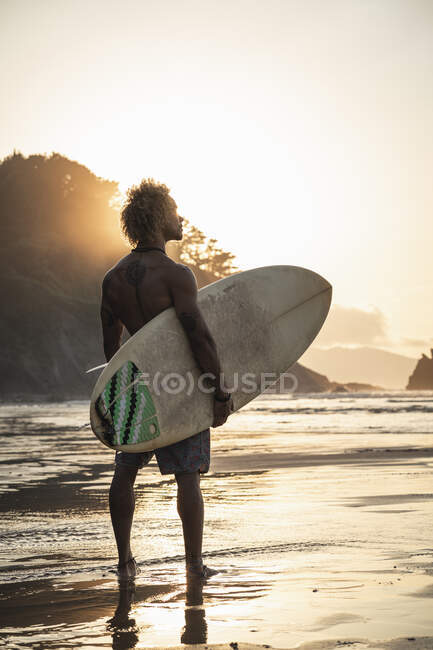 Shirtless giovane uomo con tavola da surf in piedi in riva al mare contro il cielo durante il tramonto — Foto stock