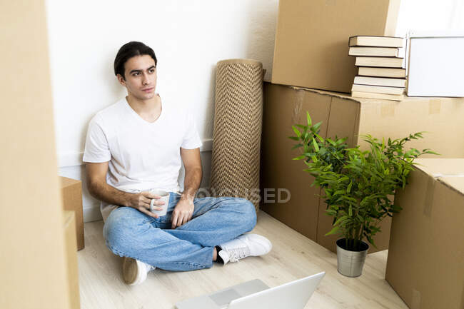 Pensativo joven sosteniendo la taza de café mientras está sentado en una casa nueva - foto de stock
