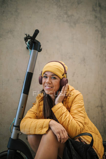 Donna sorridente che ascolta musica attraverso le cuffie mentre siede su scooter elettrico a spinta contro la parete — Foto stock