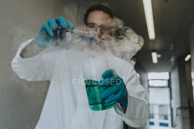Científico mezclando solución líquida en frasco mientras está de pie en el pasillo de la clínica - foto de stock