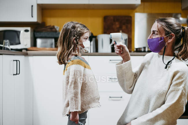 Mutter mit Gesichtsmaske überprüft Temperatur ihrer Tochter während sie zu Hause sitzt — Stockfoto