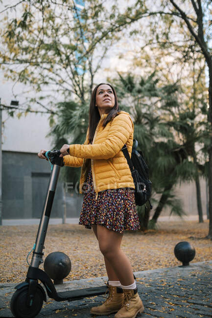 Mujer reflexiva con scooter eléctrico en la calle durante el otoño - foto de stock