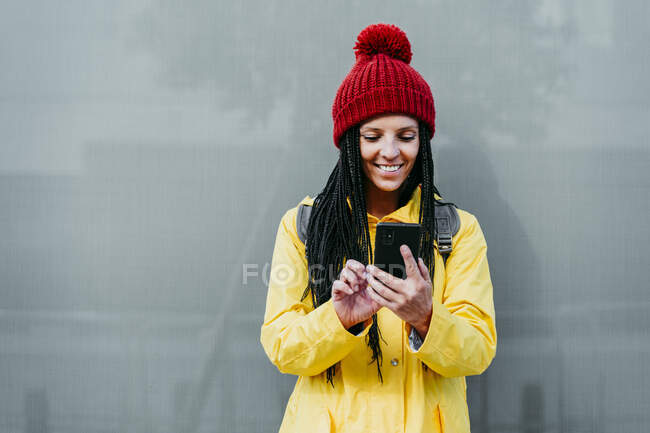 Smiling woman using smart phone standing against gray wall during autumn — Stock Photo