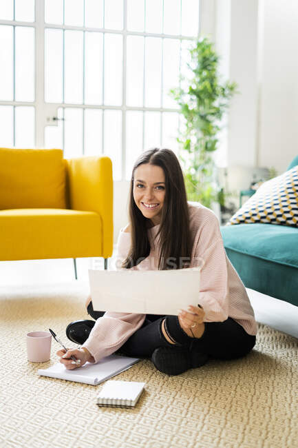 Felice bella giovane donna seduta con block notes e tazza di caffè sul tappeto contro il divano a casa — Foto stock