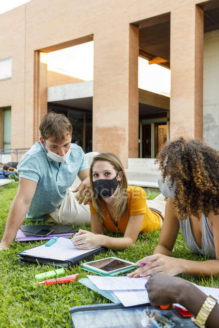 Étudiante avec des amis portant un masque de sécurité tout en étudiant sur l'herbe sur le campus universitaire — Photo de stock