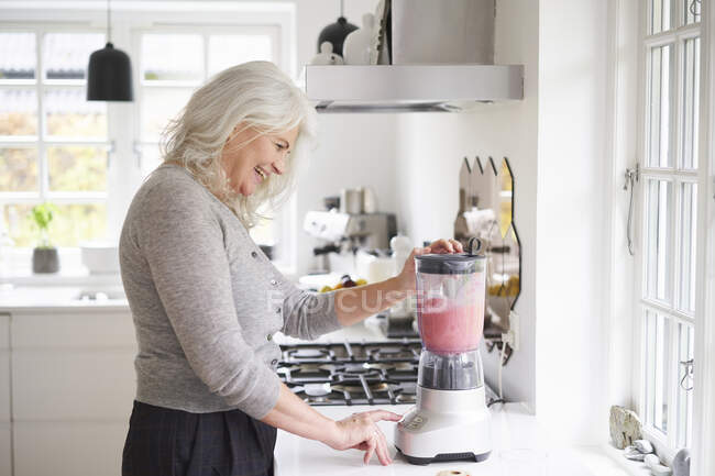 Glückliche Rentnerin bereitet Erdbeer-Smoothie in der Küchentheke zu — Stockfoto