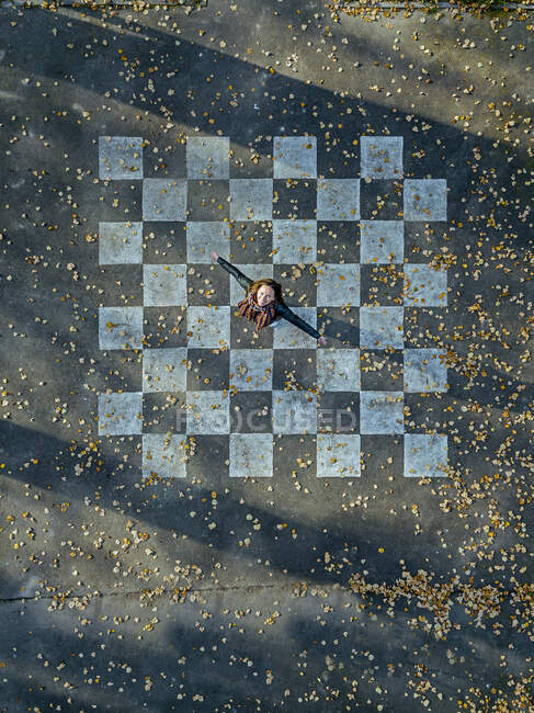 Mujer adulta con los brazos extendidos girando sobre asfalto pintado con patrón de tablero de ajedrez - foto de stock
