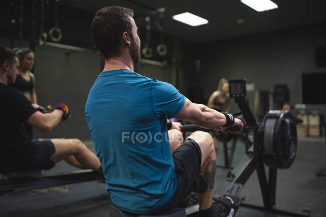 Homme utilisant la machine à ramer avec des personnes exerçant en arrière-plan à la salle de gym — Photo de stock