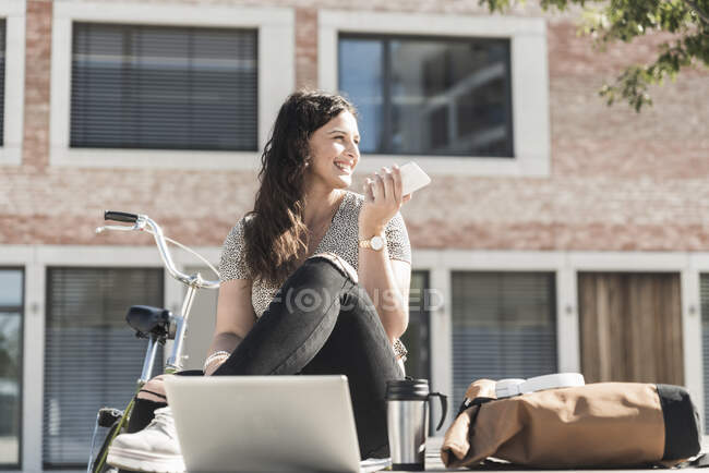 Mujer sonriente hablando por teléfono inteligente mientras se sienta contra el edificio en la ciudad — Stock Photo