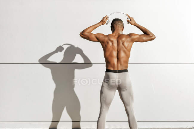 Hombre sin camisa usando auriculares mientras está de pie contra la pared blanca - foto de stock