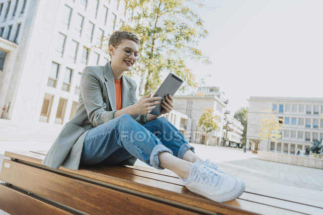 Mujer usando tableta digital mientras está sentado en el banco durante el día soleado - foto de stock