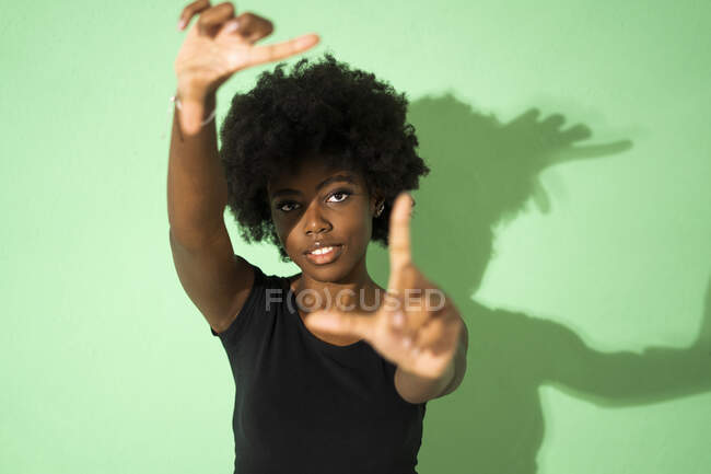 Mujer joven haciendo marco de dedo mientras está de pie sobre fondo verde - foto de stock