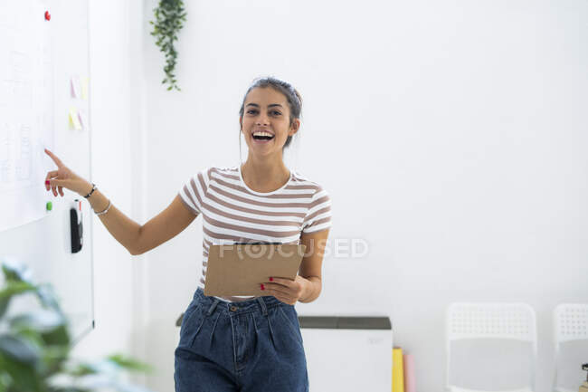 Cheerful female architect pointing at whiteboard while holding clipboard in creative workplace — Stock Photo