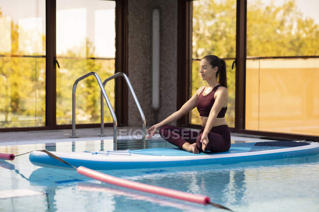 Female SUP yoga teacher meditating in lotus position over swimming pool — Stock Photo