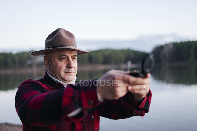 Bushcrafter con sombrero sosteniendo brújula de navegación mientras está de pie contra el lago en el bosque - foto de stock