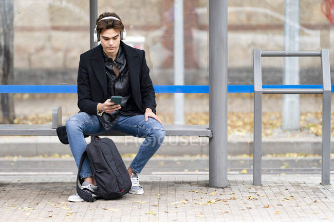 Красивый молодой человек слушает музыку, используя смартфон, сидя на скамейке на автобусной остановке — стоковое фото
