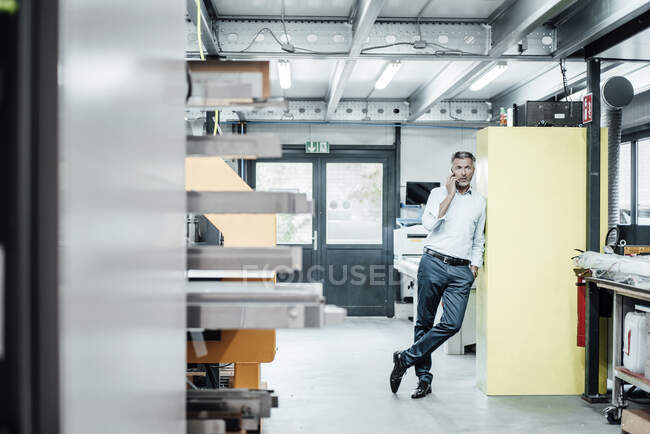 Mature businessman talking on smart phone while leaning on machinery in factory — Stock Photo