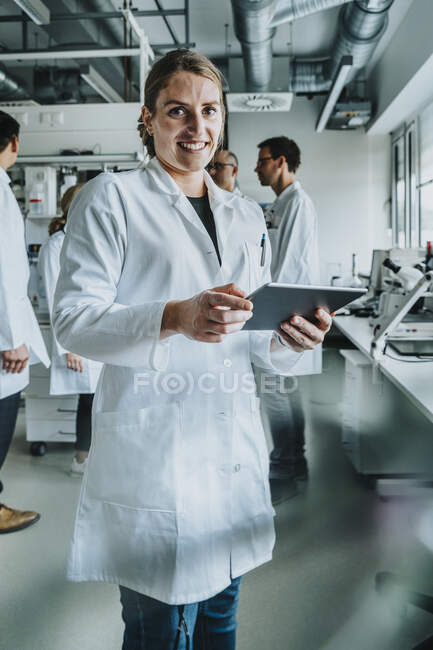 Усмішка самки за допомогою цифрового планшета, стоячи з колегою на задньому плані в лабораторії. — стокове фото