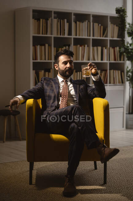 Portrait d'un homme bien habillé assis dans un fauteuil et dégustant un cigare et un verre de whisky — Photo de stock