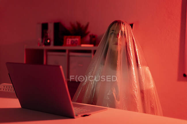 Donna premurosa coperta di plastica seduta con computer portatile alla scrivania in ufficio durante la pandemia — Foto stock