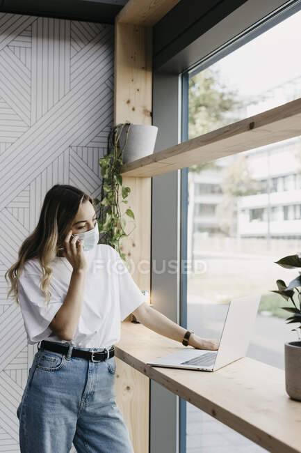 Женщина в защитной маске во время разговора на смартфоне с помощью ноутбука, стоящего в кафе во время ковида-19 — стоковое фото