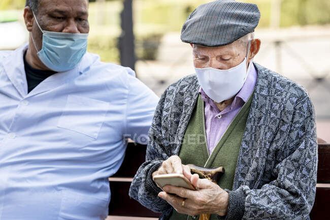 Homme âgé utilisant un téléphone intelligent portant un masque protecteur assis avec un homme mature sur un banc pendant la COVID-19 — Photo de stock