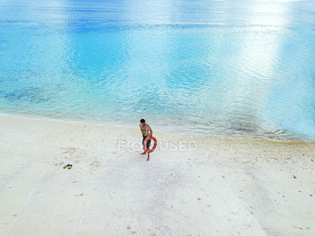 Uomo senza maglietta che tiene l'anello gonfiabile distogliendo lo sguardo mentre in piedi sulla spiaggia — Foto stock