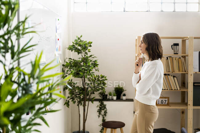 Femme d'affaires réfléchie regardant tableau blanc tout en se tenant au bureau — Photo de stock