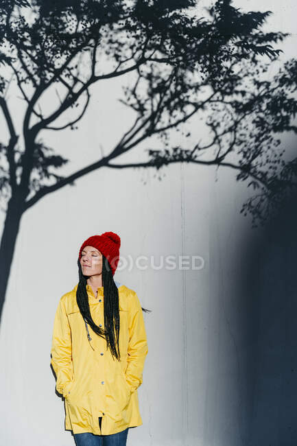 Thoughtful woman wearing raincoat standing with hands in pockets under tree shadow against wall — Stock Photo