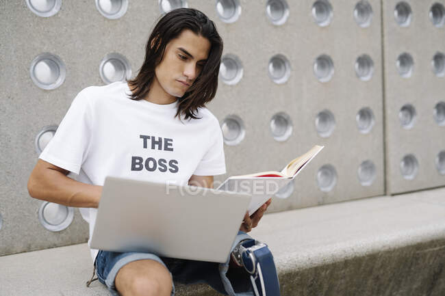 Jeune homme avec un membre artificiel travaillant sur ordinateur portable tout en étant assis contre le mur — Photo de stock
