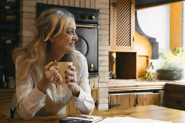 Lächelnde Frau trinkt Kaffee, während sie sich zu Hause auf Kücheninsel lehnt — Stockfoto