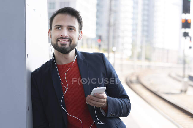 Imprenditore sorridente con cuffie in-ear in attesa sulla piattaforma ferroviaria — Foto stock