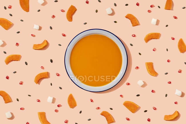 Studio shot of bowl of pumpkin soup surrounded by pumpkin seeds, pomegranate seeds and pieces of cheese and pumpkin — Stock Photo