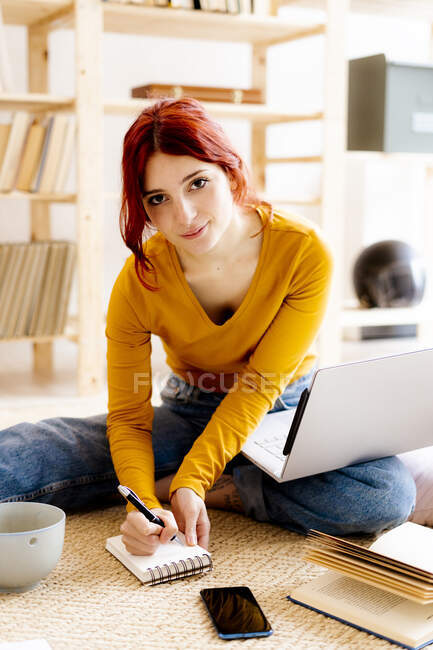 Молодая женщина с ноутбуком на коленях, записывая в блокнот во время учебы дома — стоковое фото