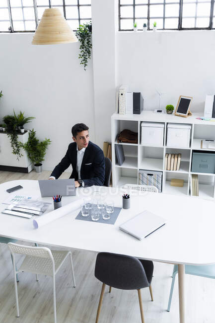 Nachdenklicher männlicher Jungunternehmer schaut weg, während er mit Laptop am Schreibtisch am Arbeitsplatz sitzt — Stockfoto