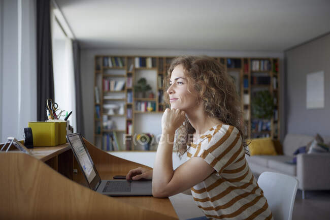 Lächelnde Frau mit Kopf in den Händen sitzt zu Hause am Schreibtisch — Stockfoto
