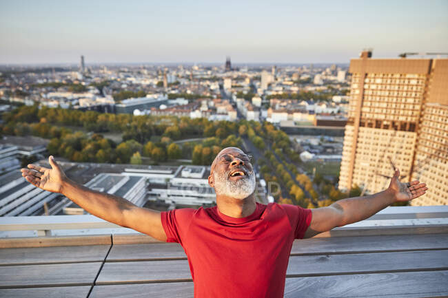 Hombre barbudo feliz con los brazos extendidos inclinados hacia atrás mientras está de pie en la terraza del edificio en la ciudad durante la puesta del sol - foto de stock