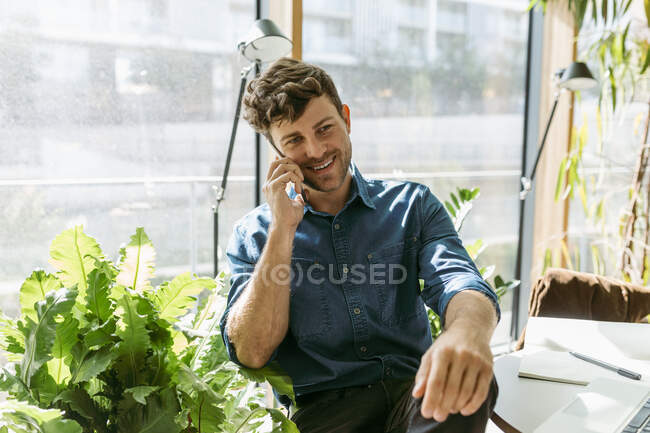 Sonriente joven empresario hablando por teléfono inteligente mientras está sentado en la cafetería - foto de stock
