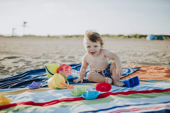 Mignon petit garçon jouant tout en étant assis sur une serviette colorée à la plage pendant le coucher du soleil — Photo de stock