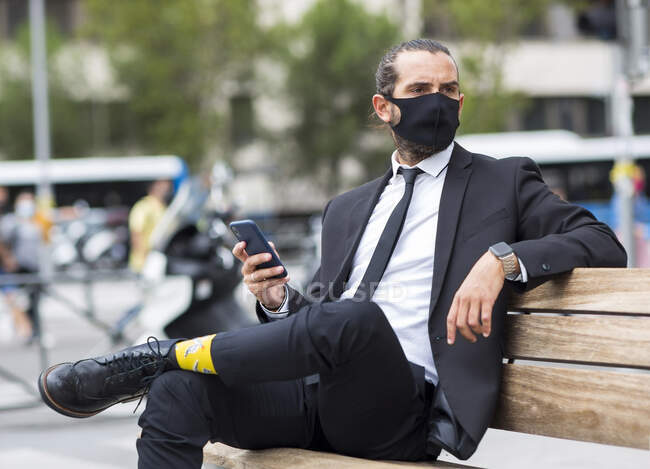Портрет бизнесмена в защитной маске, сидящего на скамейке с смартфоном в руке — стоковое фото