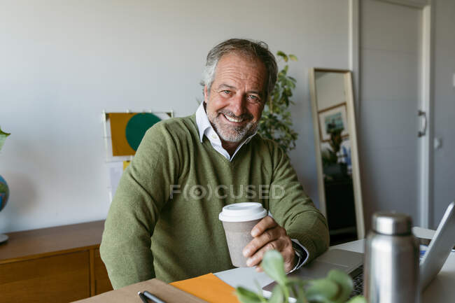 Sonriente hombre maduro sosteniendo taza de café desechable sentado en casa - foto de stock