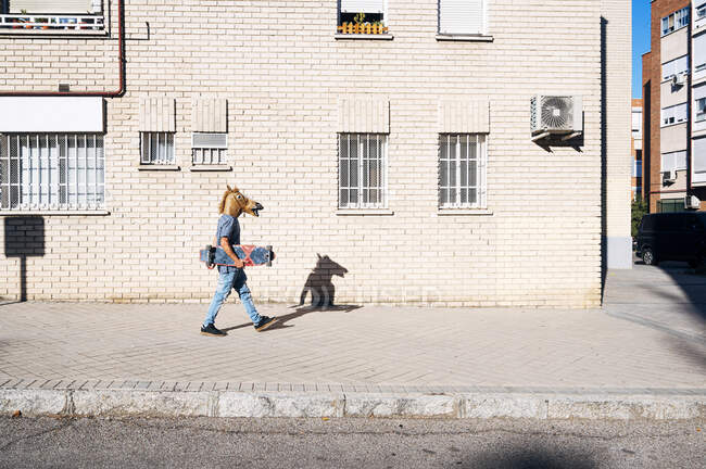Uomo in maschera di cavallo a piedi con skateboard sul sentiero costruendo in città durante la giornata di sole — Foto stock
